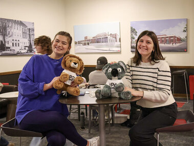 Two people seated at a table holding stuffed animals in a room with framed architectural art on walls. Photo by Nicole Gibson.